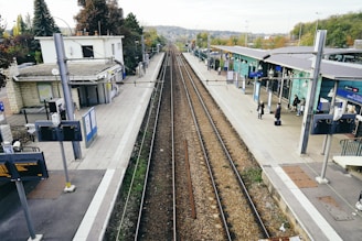 A railway station platform featuring train tracks running down the center, bordered by platforms on either side. The area appears relatively quiet with a few people waiting. Buildings and signage typical of a train station are visible, along with a small number of trees and greenery in the background.