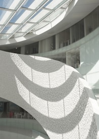 A modern architectural interior featuring a curved, textured wall illuminated by sunlight streaming through a glass ceiling. The shadows cast intricate patterns on the wall's surface, enhancing the contemporary design. The space appears open and airy, with glass railings and minimalist decor in the background.