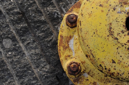 A weathered and rusted yellow cap or flange, held in place by four rusty bolts. The surface has a textured, gritty appearance with visible signs of wear and corrosion. It is positioned against a gray textured background that seems to be a concrete surface.