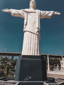 A large statue resembling the Christ the Redeemer, with arms outstretched, stands prominently against a clear blue sky. The statue is mounted on a dark pedestal, and there is a bridge in the background with some trees and other structures nearby.