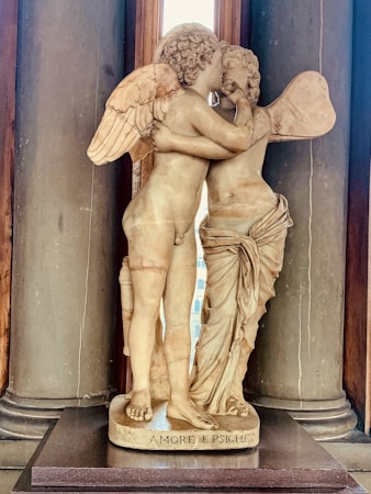 A classical marble sculpture of two winged figures, embracing intimately in a tender kiss. The figures are adorned with detailed wings and are partly clothed, positioned in an elegant pose that captures a moment of affection.