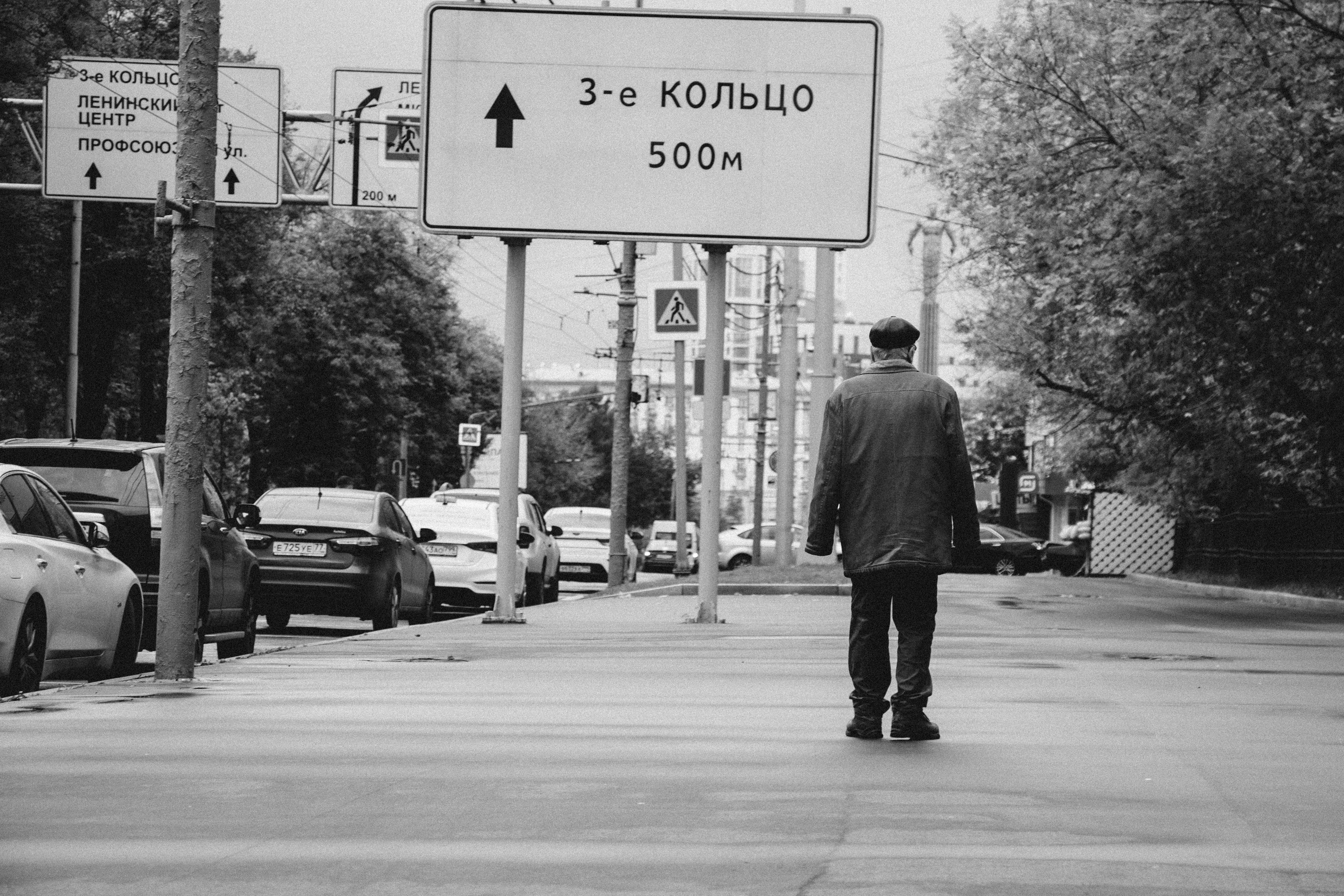 grayscale photography of man walking on sidewalk beside roadsign