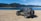 A rugged black Jeep is parked on a sandy terrain beside a large, tranquil lake. In the background, dense forest-covered mountains rise under a clear blue sky.