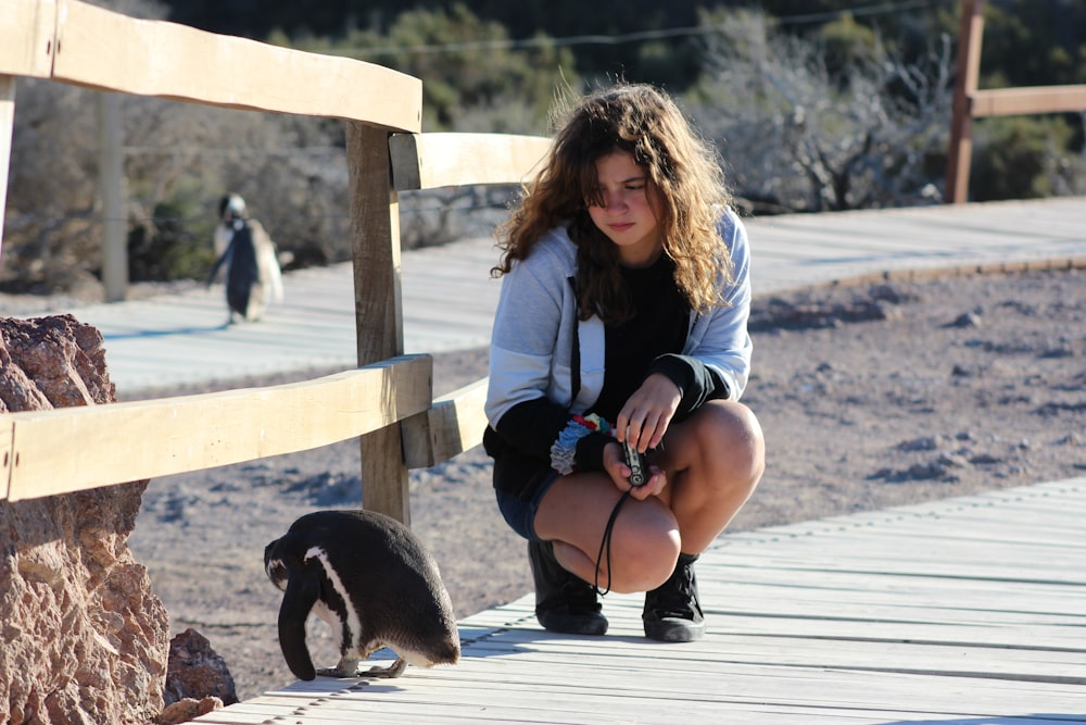 woman wearing white and black jacket sitting on wooden dock near penguin during daytime