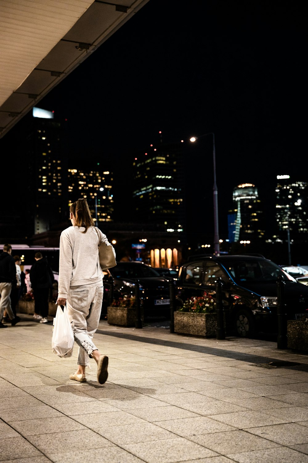 woman wearing white long-sleeved shirt holding plastic bag walking on pathway during night time