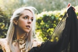 A woman with long, light hair styled with small braids gazes thoughtfully into the distance. She wears a garment adorned with gold sequins and holds a black, sheer fabric that flows gracefully from her shoulder. The background is a lush, green, natural setting, softly blurred, suggesting a garden or park.