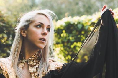 A woman with long, light hair styled with small braids gazes thoughtfully into the distance. She wears a garment adorned with gold sequins and holds a black, sheer fabric that flows gracefully from her shoulder. The background is a lush, green, natural setting, softly blurred, suggesting a garden or park.