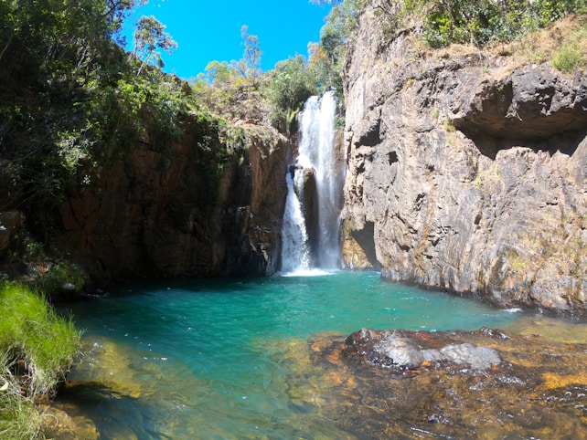 A scenic waterfall cascades down a rugged cliff into a tranquil, turquoise pool surrounded by lush green foliage. Sunlight illuminates the water and rocks, creating a natural and serene setting.