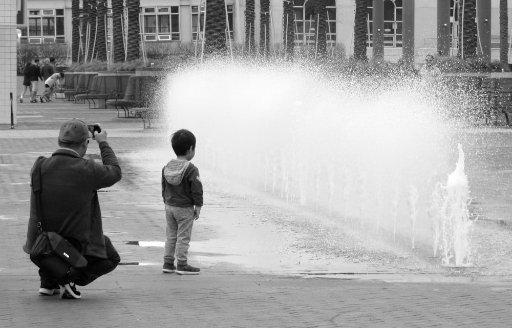 fotografia in scala di grigi di persona che scatta foto di bambino in piedi vicino alla fontana d'acqua