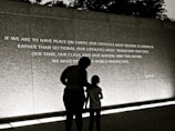 A large wall displays an inspirational quote, with two individuals silhouetted against it. The quote calls for peace and broader perspectives beyond societal divisions. The scene is captured in low light or a sepia tone, creating a contemplative atmosphere.