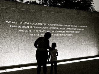 A large wall displays an inspirational quote, with two individuals silhouetted against it. The quote calls for peace and broader perspectives beyond societal divisions. The scene is captured in low light or a sepia tone, creating a contemplative atmosphere.
