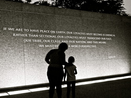 A large wall displays an inspirational quote, with two individuals silhouetted against it. The quote calls for peace and broader perspectives beyond societal divisions. The scene is captured in low light or a sepia tone, creating a contemplative atmosphere.