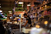 A bustling market scene features a vendor at a counter surrounded by various jars, bottles, and packaged goods on shelves. The vendor is focused on serving customers while a group of people stands nearby. Ambient lighting adds warmth to the crowded and vibrant environment.