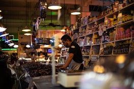 A bustling market scene features a vendor at a counter surrounded by various jars, bottles, and packaged goods on shelves. The vendor is focused on serving customers while a group of people stands nearby. Ambient lighting adds warmth to the crowded and vibrant environment.