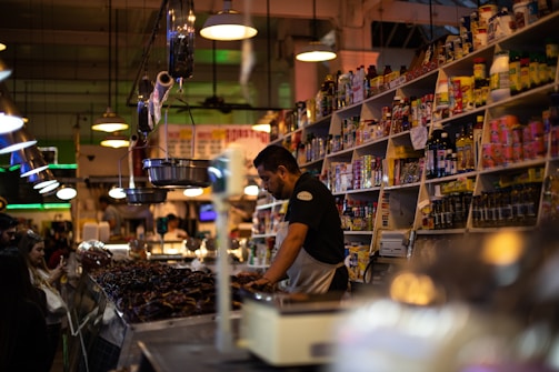 A bustling market scene features a vendor at a counter surrounded by various jars, bottles, and packaged goods on shelves. The vendor is focused on serving customers while a group of people stands nearby. Ambient lighting adds warmth to the crowded and vibrant environment.
