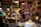A bustling market scene features a vendor at a counter surrounded by various jars, bottles, and packaged goods on shelves. The vendor is focused on serving customers while a group of people stands nearby. Ambient lighting adds warmth to the crowded and vibrant environment.