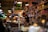 A bustling market scene features a vendor at a counter surrounded by various jars, bottles, and packaged goods on shelves. The vendor is focused on serving customers while a group of people stands nearby. Ambient lighting adds warmth to the crowded and vibrant environment.