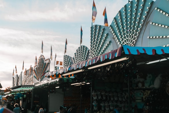 A vibrant carnival scene with brightly lit signs and decorative elements lining the top of stalls. The architecture includes triangular light patterns and flags, suggesting a festive atmosphere. Below, numerous stalls are adorned with prizes, such as stuffed animals. People are seen walking, adding to the lively environment.