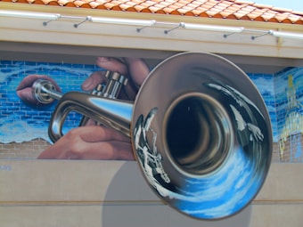 A mural depicting a large, shiny trumpet being played by hands. The background features a blue sky painted on a brick wall. The image is artistic and detailed.