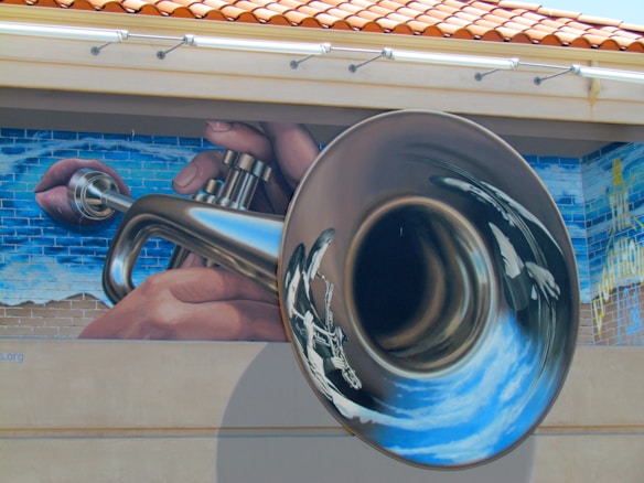 A mural depicting a large, shiny trumpet being played by hands. The background features a blue sky painted on a brick wall. The image is artistic and detailed.