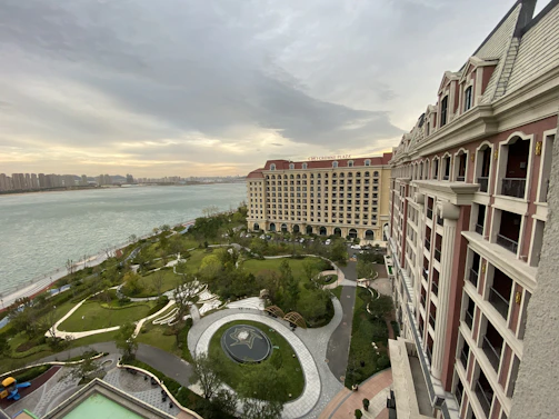 An aerial view of a well-kept garden area with winding pathways and a circular water feature, located by the side of a wide river. In the background, there is a large, elegant hotel building with multiple floors and a distinctive red roof. The skyline on the opposite side of the river shows a series of high-rise buildings under a cloudy sky.