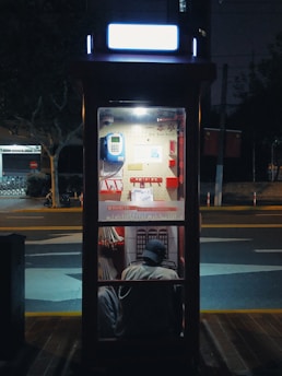 A dimly lit phone booth on a street at night, featuring a blue phone mounted on the interior wall with a small chair and a person sitting inside wearing a cap.