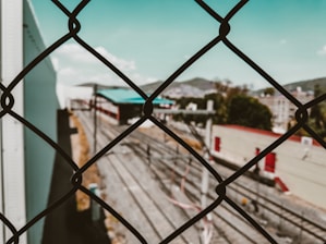 A chain-link fence in the foreground with a blurred view of train tracks and industrial buildings in the background. The sky is clear with a few clouds, and there are hints of greenery beyond the tracks.