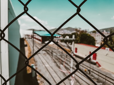 A chain-link fence in the foreground with a blurred view of train tracks and industrial buildings in the background. The sky is clear with a few clouds, and there are hints of greenery beyond the tracks.