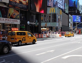 A busy urban street scene featuring yellow taxis, a crowd of people, and prominent retail signage. Large digital billboards and advertisements are displayed on the surrounding buildings. The scene conveys the hustle and bustle of a vibrant city environment.