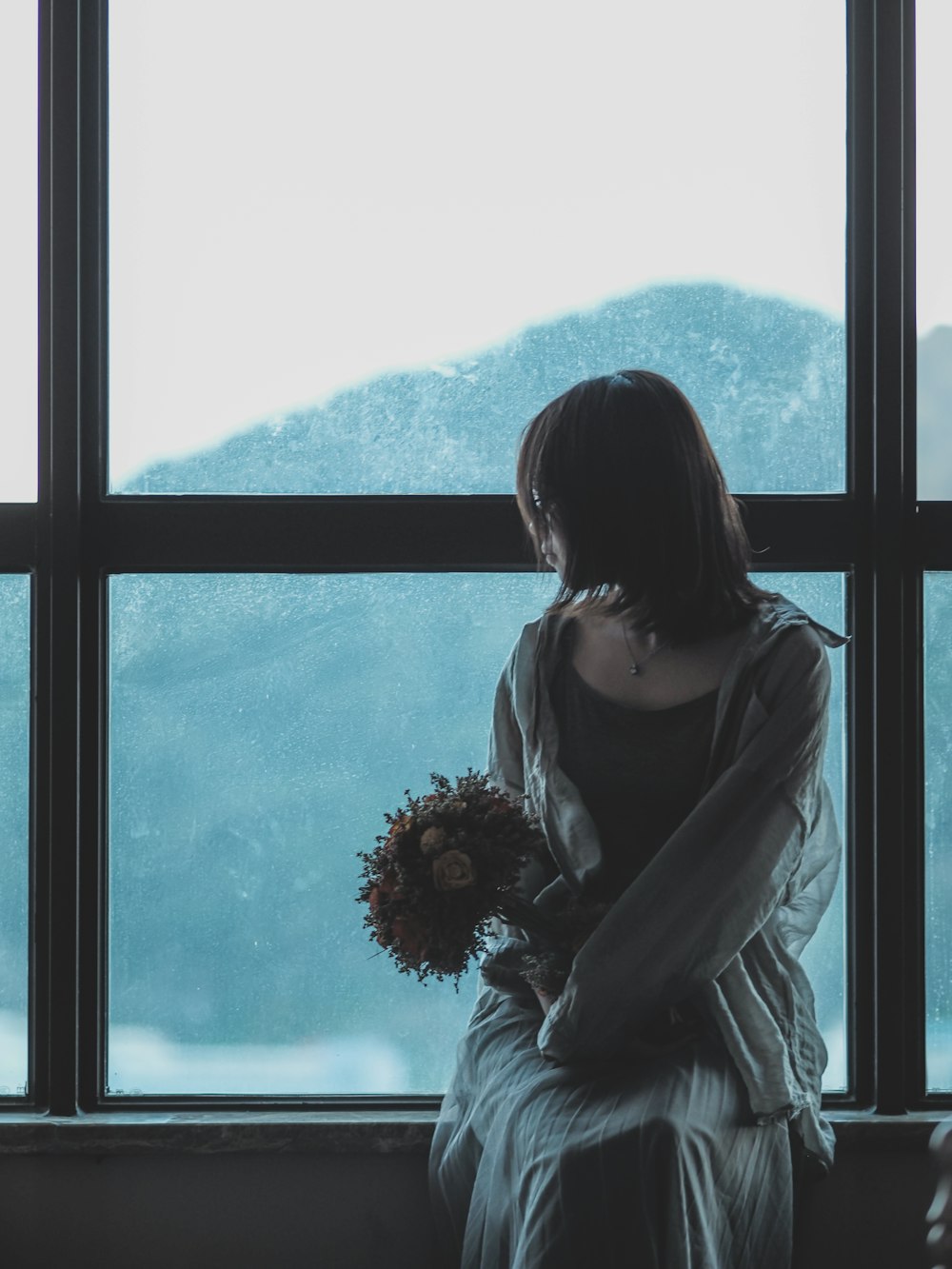 Frau sitzt an einem Glasfenster und hält einen Blumenstrauß in der Hand