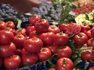 A vibrant display of fresh red tomatoes stacked on a market stand, surrounded by green leaves. In the background, there are other fruits and vegetables, including blueberries and a blurred variety of produce. A sliced tomato is visible among the whole ones, showcasing its juicy interior. A hand reaches from the top left corner, possibly selecting a tomato.