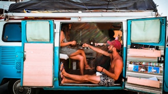 A couple is sitting inside a retro-style camper van with open doors, enjoying a relaxed moment. The woman is seated on a stool while the man sits on the floor barefoot, both engaged in conversation or sharing a drink. The interior of the van is equipped with various stored items and decorations, creating a cozy and personalized space. The van exterior is painted turquoise with black and brown highlights, adding a vibrant touch to the scene.