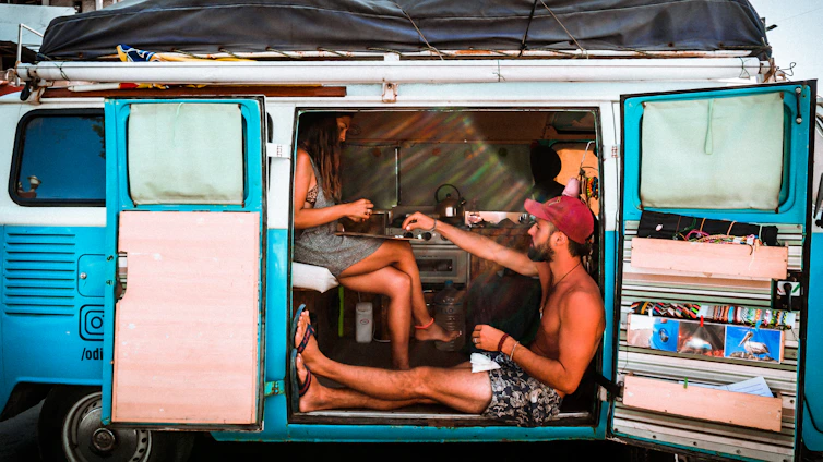 A happy couple working on laptops inside a cozy campervan overlooking a serene Australian beach at sunset.