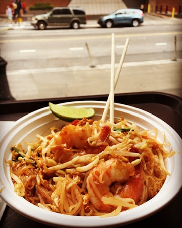A bowl of pad Thai noodles garnished with shrimp, bean sprouts, and a lime wedge sits on a table. Chopsticks rest on the bowl. The scene is set near a window overlooking a street with passing cars and pedestrians.