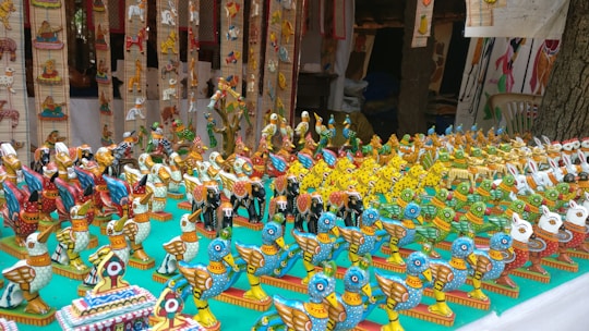 A display of colorful, handcrafted wooden bird figurines arranged on a table. These figurines are intricately painted in bright hues with patterns, showcasing a variety of bird designs. The setup is likely part of a market stall or exhibition, with more decorations visible in the background.