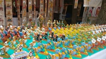 A display of colorful, handcrafted wooden bird figurines arranged on a table. These figurines are intricately painted in bright hues with patterns, showcasing a variety of bird designs. The setup is likely part of a market stall or exhibition, with more decorations visible in the background.
