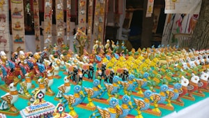 A display of colorful, handcrafted wooden bird figurines arranged on a table. These figurines are intricately painted in bright hues with patterns, showcasing a variety of bird designs. The setup is likely part of a market stall or exhibition, with more decorations visible in the background.