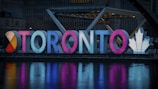Illuminated letters spelling 'Toronto' are displayed in a vibrant design. The letters are brightly lit in various colors, including red, blue, and pink. A large, white maple leaf symbol is featured on the right side, and the scene is reflected in a body of water in the foreground.