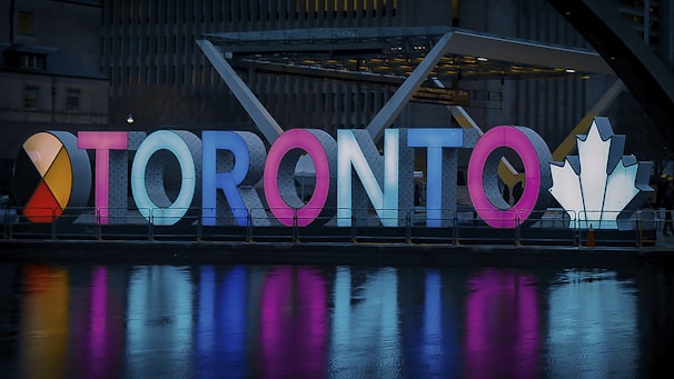 Illuminated letters spelling 'Toronto' are displayed in a vibrant design. The letters are brightly lit in various colors, including red, blue, and pink. A large, white maple leaf symbol is featured on the right side, and the scene is reflected in a body of water in the foreground.