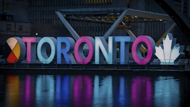 Illuminated letters spelling 'Toronto' are displayed in a vibrant design. The letters are brightly lit in various colors, including red, blue, and pink. A large, white maple leaf symbol is featured on the right side, and the scene is reflected in a body of water in the foreground.