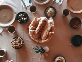 A dining table set with a variety of dishes. In the center, there is a basket filled with flatbreads. Surrounding it are bowls containing different types of food including salad, couscous, and sauces. The table is covered with a beige tablecloth and includes some empty plates, cups, and cutlery.