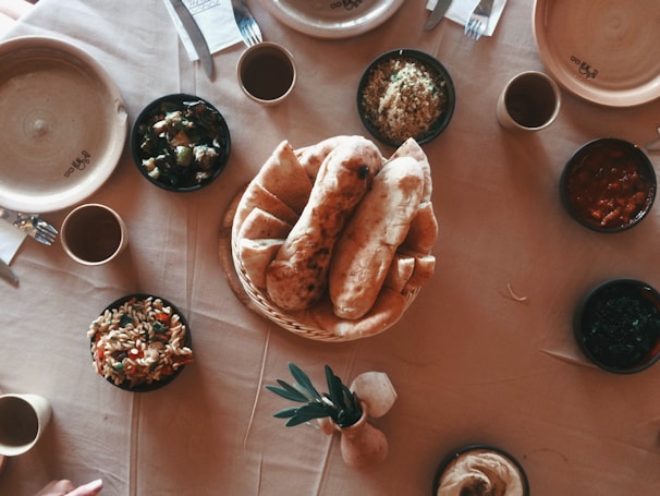 A dining table set with a variety of dishes. In the center, there is a basket filled with flatbreads. Surrounding it are bowls containing different types of food including salad, couscous, and sauces. The table is covered with a beige tablecloth and includes some empty plates, cups, and cutlery.