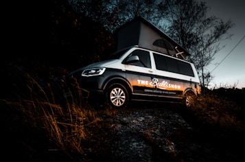 A parked van on a grassy hill at dusk with an elevated roof, partially surrounded by trees. The lighting creates a dramatic contrast, highlighting the text on the van's side.