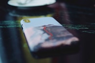 A close-up of a smartphone placed on a reflective, dark surface. The phone has a case featuring a vibrant image of a mountain with a red path or lava flow. The background is blurred, focusing attention on the phone case design.