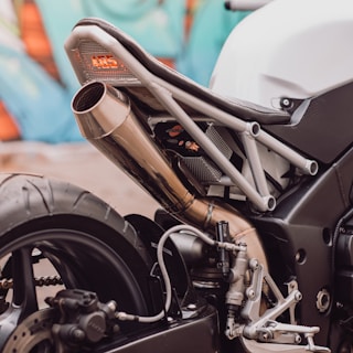 A close-up view of the rear section of a motorcycle featuring the exhaust pipe, part of the frame, and the rear wheel. The metal exhaust has a polished finish, and the frame has a modern, industrial design. The background showcases abstract, colorful graffiti.