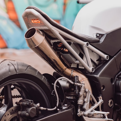 A close-up view of the rear section of a motorcycle featuring the exhaust pipe, part of the frame, and the rear wheel. The metal exhaust has a polished finish, and the frame has a modern, industrial design. The background showcases abstract, colorful graffiti.