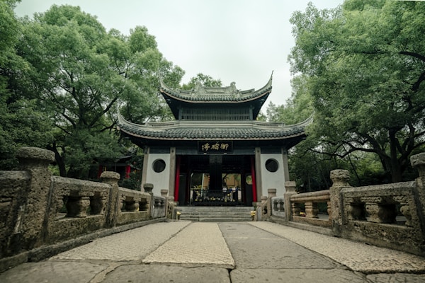 A traditional Chinese pavilion is surrounded by lush green trees, with a stone pathway leading up to it. The pavilion has a graceful curved roof and wooden details, conveying an atmosphere of tranquility.