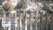 A wooden fence with vertical posts partially obstructs the view of a serene lakeside setting. A sign in multiple languages indicates that swimming and fishing are forbidden. Behind the fence, the lake is surrounded by trees and rock formations.