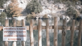 A wooden fence with vertical posts partially obstructs the view of a serene lakeside setting. A sign in multiple languages indicates that swimming and fishing are forbidden. Behind the fence, the lake is surrounded by trees and rock formations.