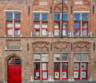 A brick facade of an old building with red-framed windows and intricate stone detailing. The architecture features symmetrical windows with decorative stone arches and a prominent red door at the bottom.
